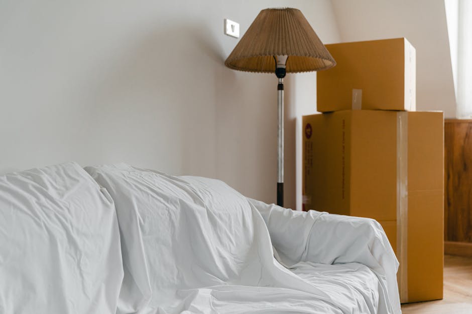 Inside a home during a move, a white sofa covered with a protective cloth is positioned in front of a light-colored wall. Behind the sofa, there is a tall, freestanding lamp with a chestnut brown lamp