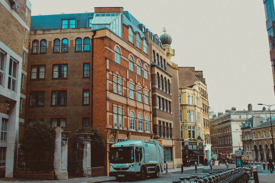 A white removal truck from Maida Hill Removals is parked on a street curb outside a multi-storey building with a brick facade and large windows, where furniture and packing boxes are visible on the pa