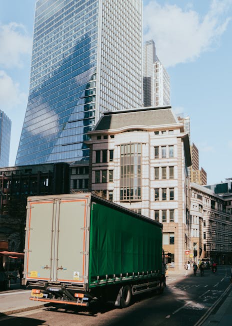 A white removal truck from Maida Hill Removals is parked on a street curb outside a multi-storey building with a brick facade and large windows, where furniture and packing boxes are visible on the pa